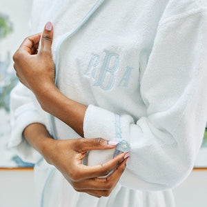 A model wears a white bath robe with light blue piping and monogram. She buttons the cuff of her sleeve.