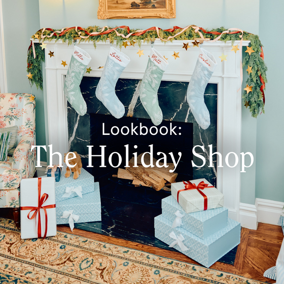 Holiday-themed fireplace mantel decorated with garland, ribbon, stars, personalized stockings, and wrapped gifts beneath “The Holiday Shop” text.