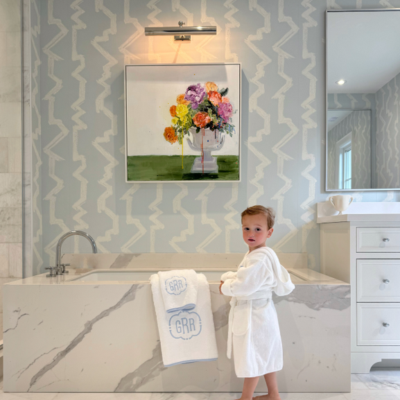 Child in a white robe standing beside a marble bathtub with monogrammed towels in an elegant blue-and-white bathroom.