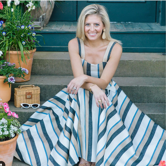 Woman in striped blue-and-white dress sitting on front steps beside flowers and wicker basket, smiling at the camera outdoors.