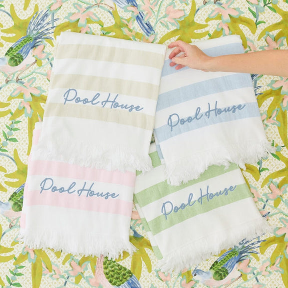 Four striped beach towels embroidered with “Pool House” arranged on a colorful tropical-patterned fabric backdrop.