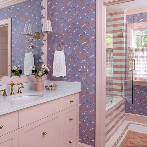 Pink bathroom vanity with brass fixtures, lavender floral wallpaper, scalloped mirror trim, and striped pink tile shower.