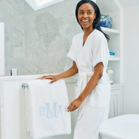 A woman wearing a sage green Palmero patterned robe holds open a matching bath towel.