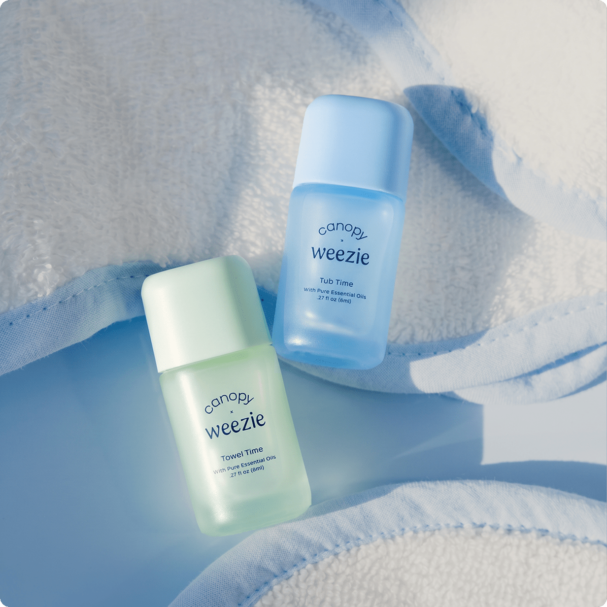 Two small Canopy Weezie Towel Time travel size bottles, one in light mint green and one in light sky blue, arranged on a soft white towel with blue shadows.