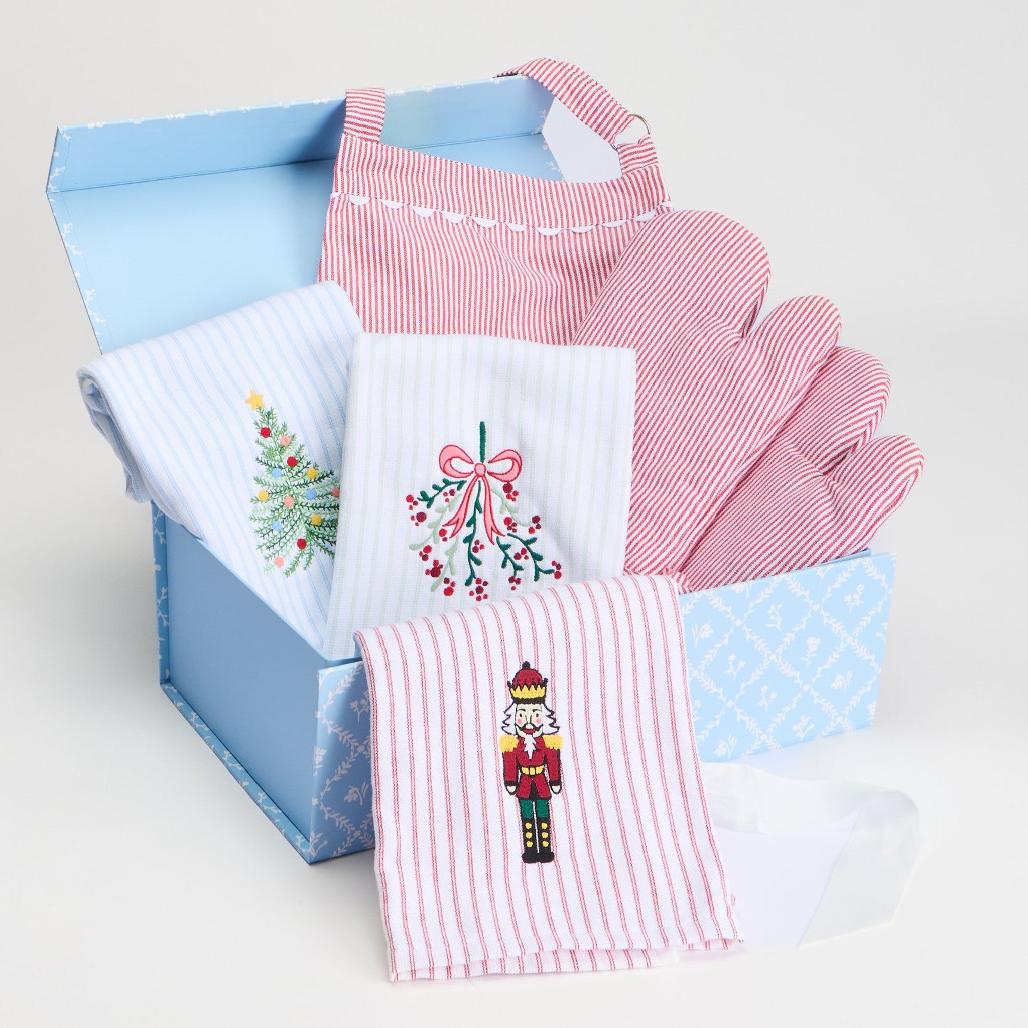 Blue gift box with striped hand towels embroidered with a Christmas tree, mistletoe and nutcracker, plus a red striped apron.