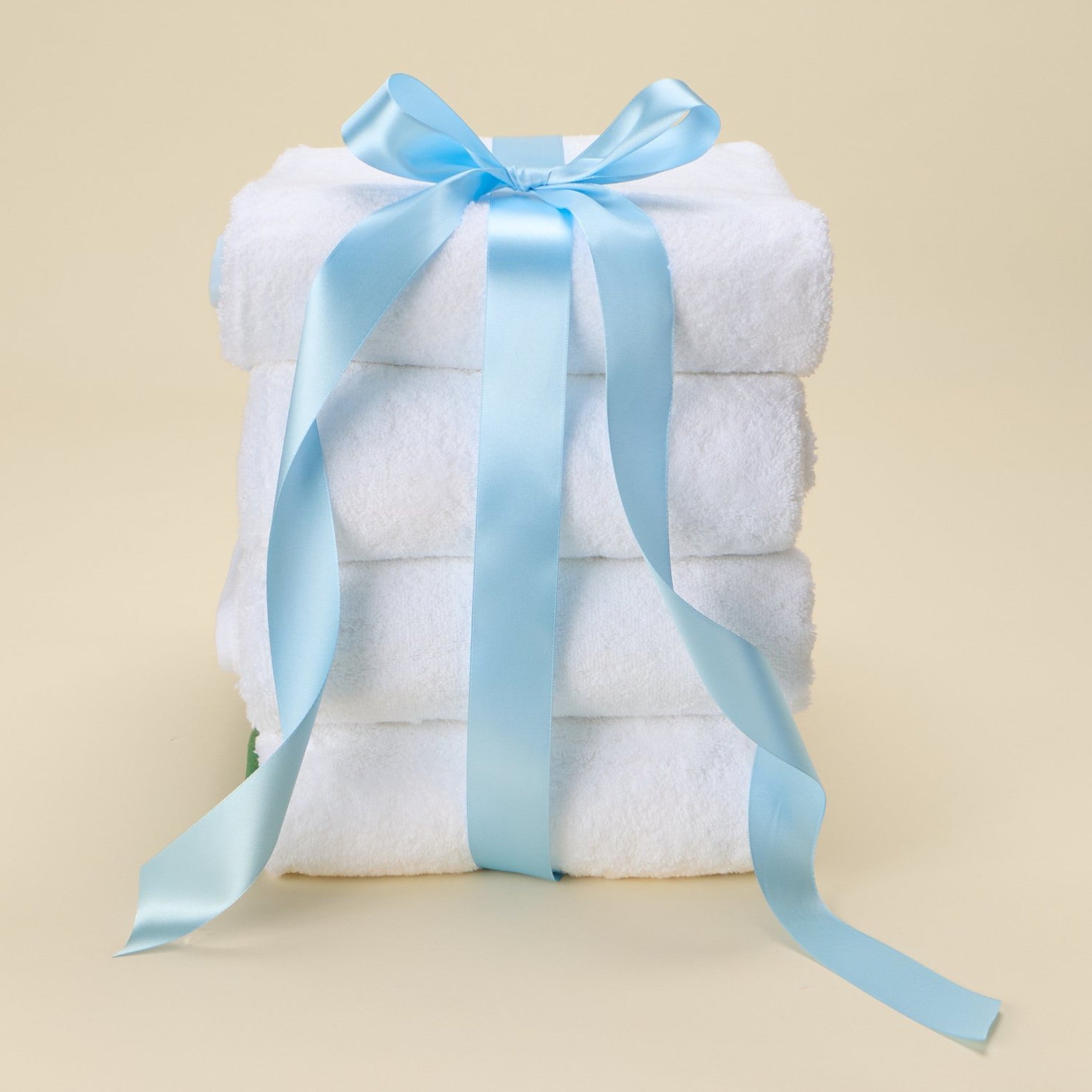 Stack of folded white Weezie bath towels tied with a light blue satin ribbon bow on a beige background.