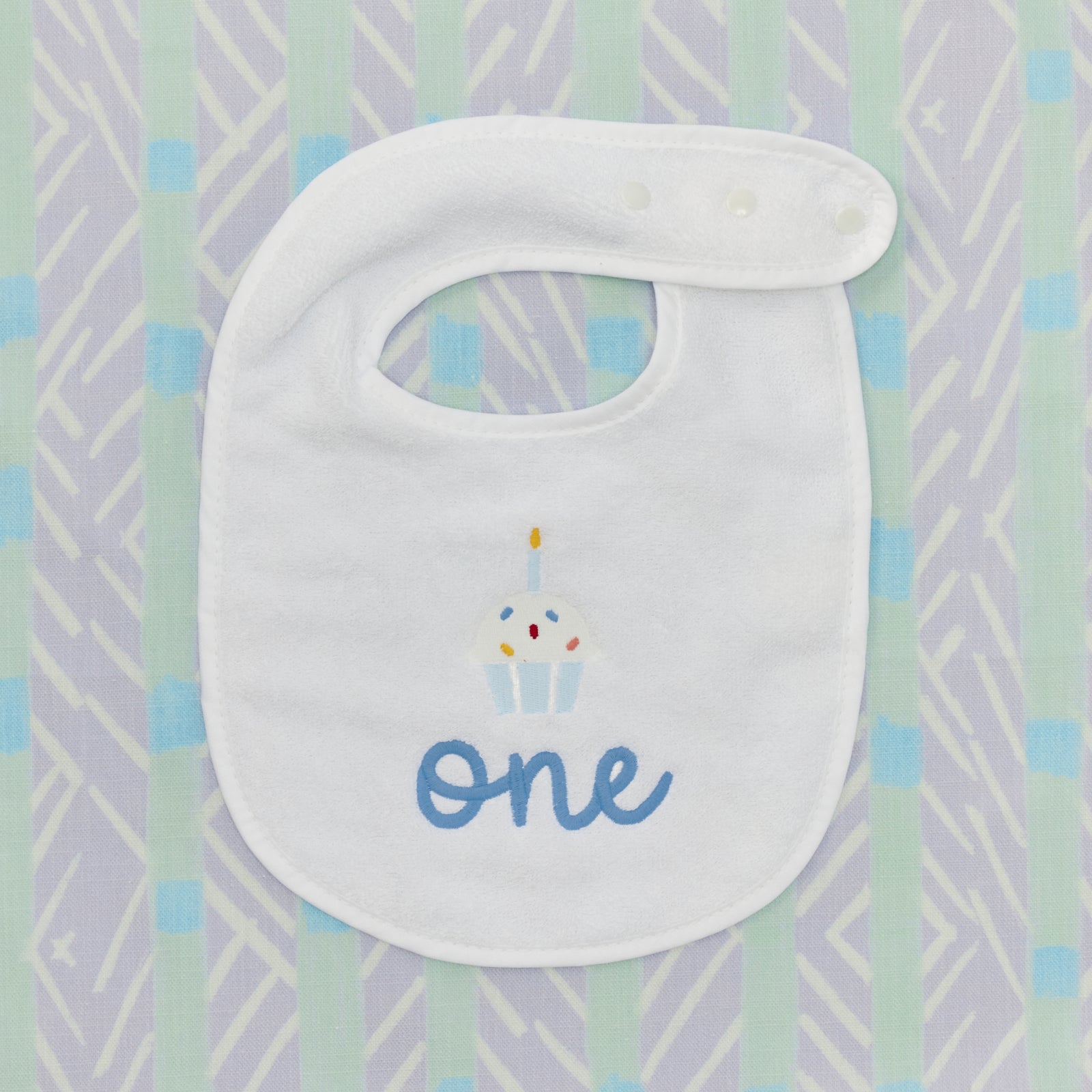 Detail of the embroidered cupcake and blue 'one' on a white birthday bib.