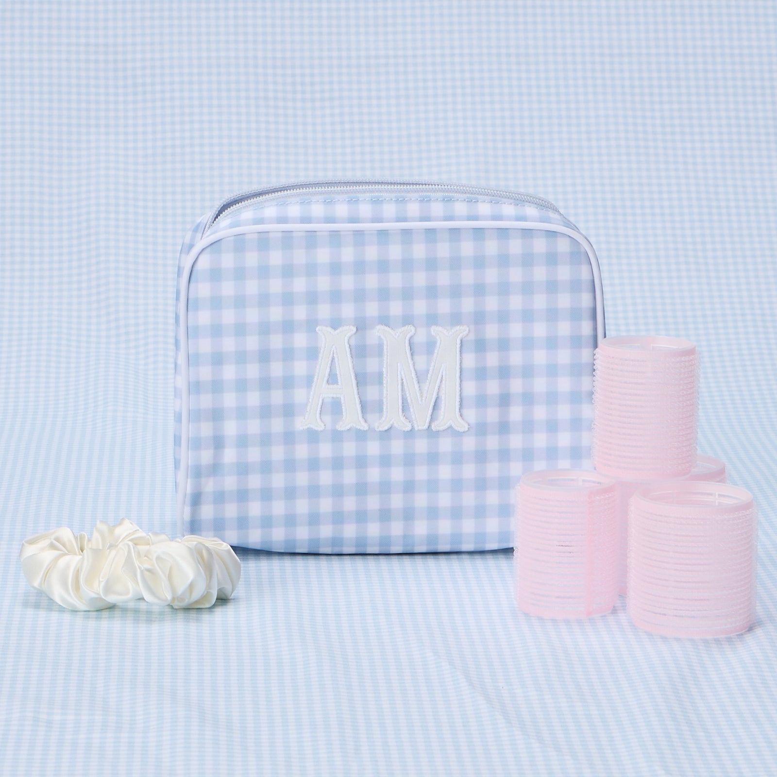 A medium, light blue gingham patterned toiletry bag with "AM" embroidered in white, sitting next to pink hair rollers.