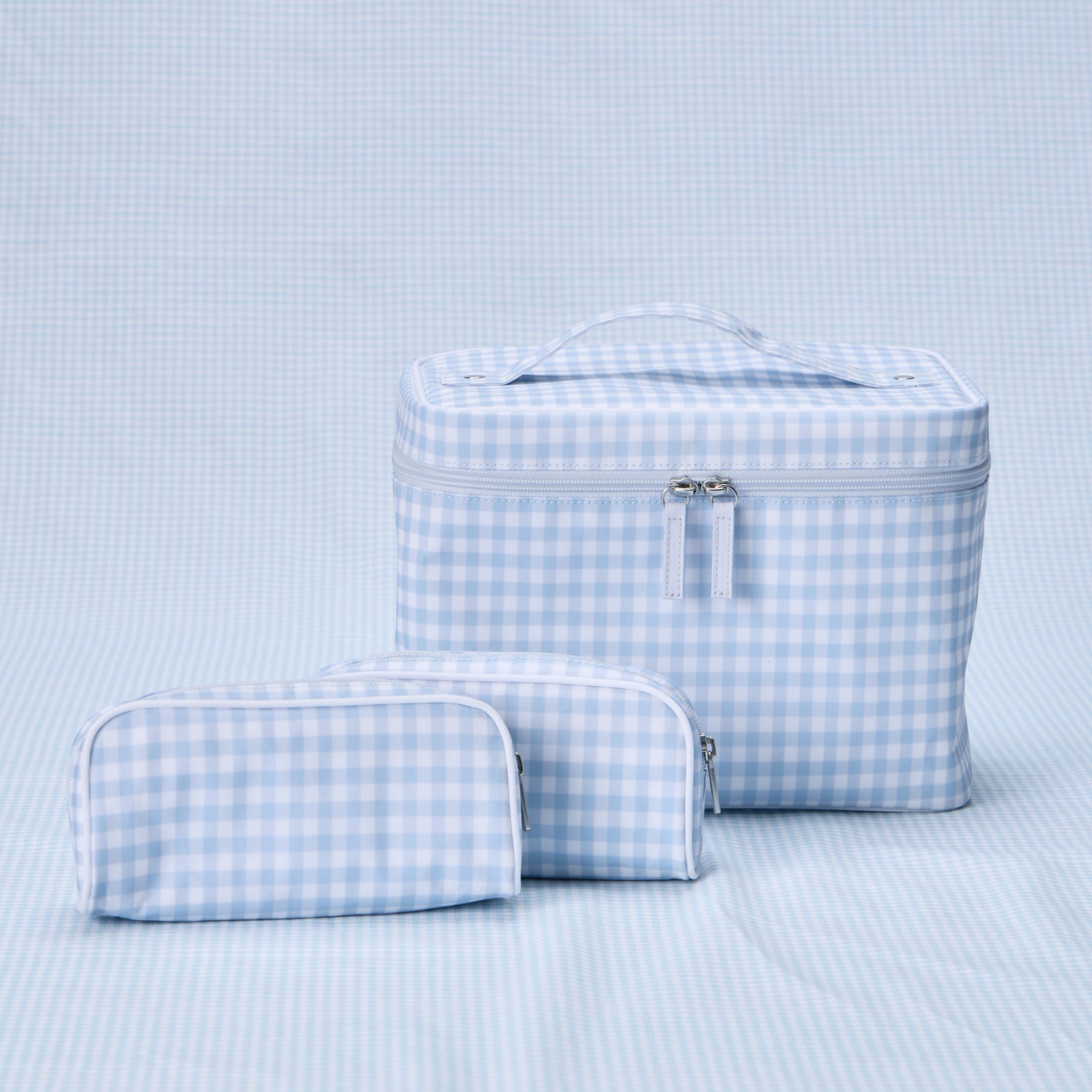 A Weezie Travel Bundle in Light Blue Gingham, consisting of one large, structured vanity case with a top handle and two small toiletry bags, all featuring a matching blue and white checkered pattern.