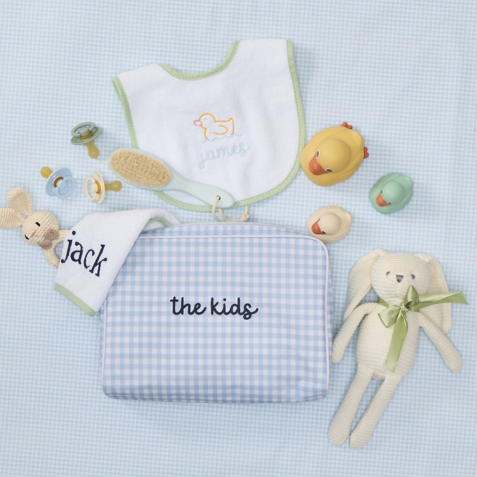 Light blue gingham toiletry bag embroidered with "the kids," next to a baby bib, pacifiers, a brush, and two toy bunnies.