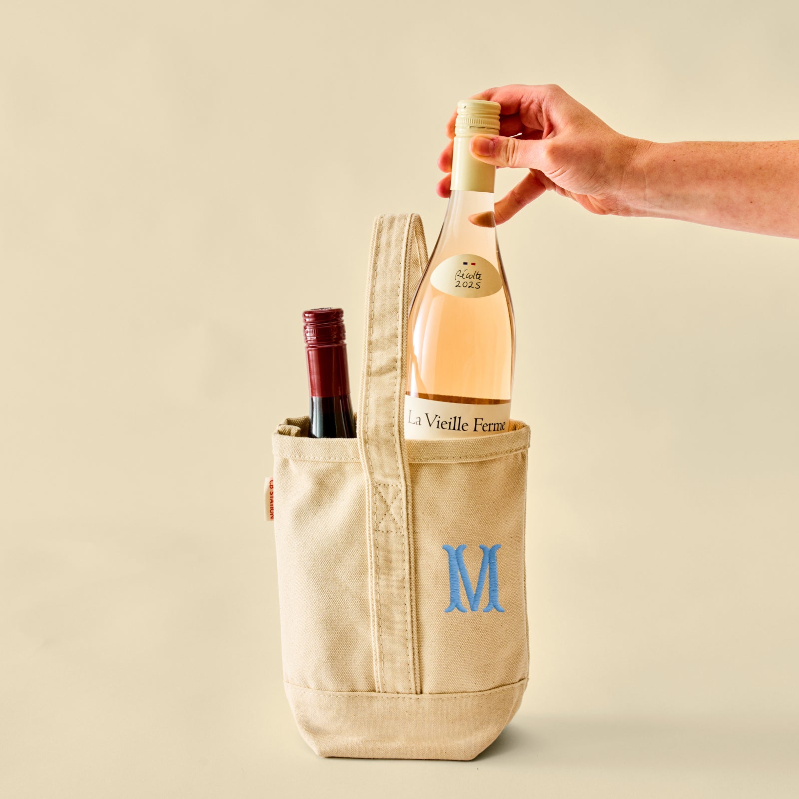 A beige canvas wine tote bag with a blue embroidered letter 'M' on the front pocket, holding a bottle of rosé wine and another bottle in a side pocket, with a hand reaching in to lift the top bottle. Photographed against a solid beige background.