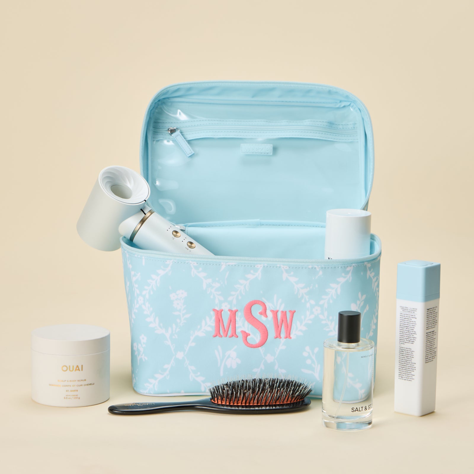 An open, light blue vanity case with a white trellis floral pattern, featuring a custom pink 'MSW' monogram embroidered on the front. The case is packed with beauty essentials, including a hair dryer, a jar of OUAI scrub, a hairbrush, and other skincare bottles.