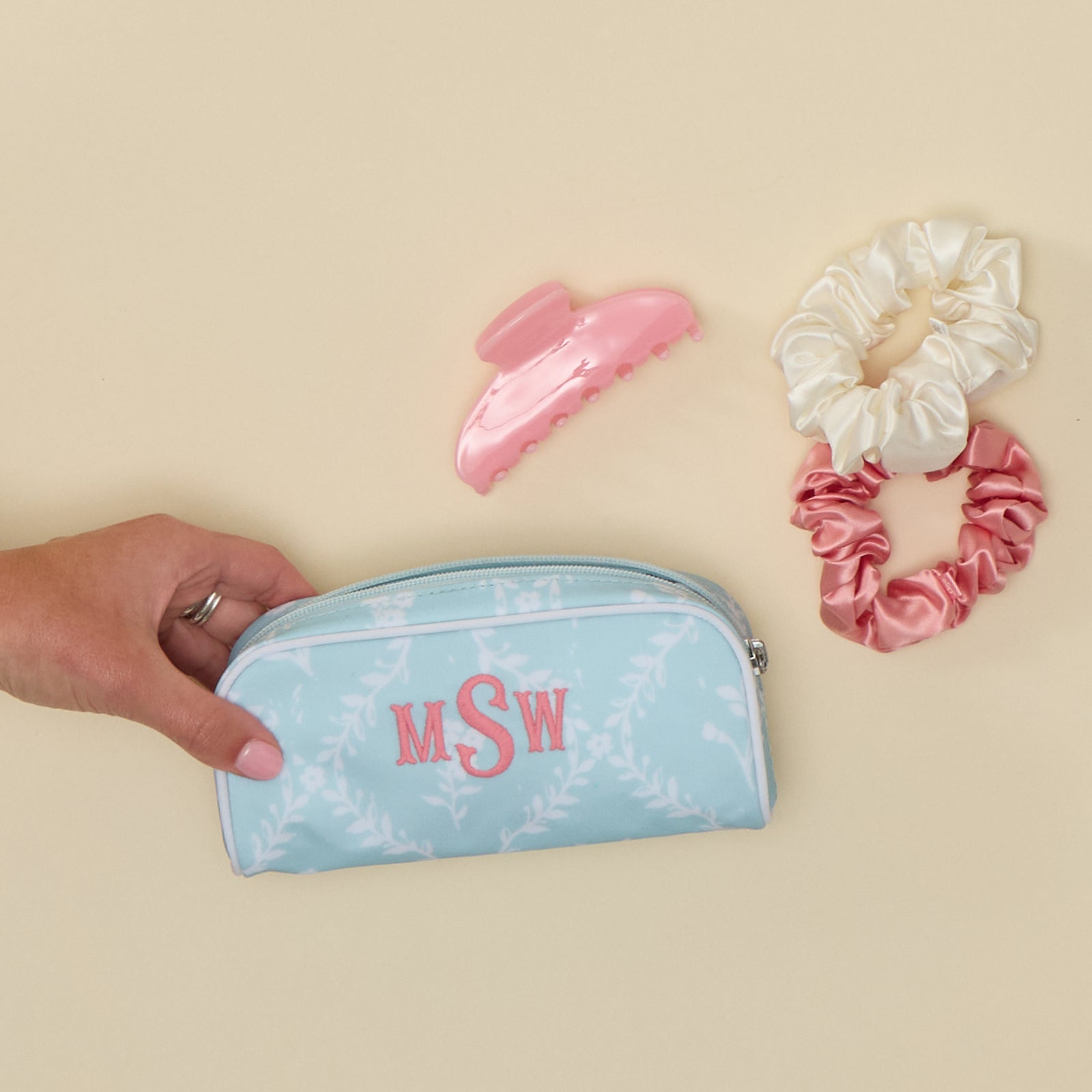 Hand holding a small blue trellis toiletry bag with "MSW" embroidery next to a pink hair clip and two scrunchies.
