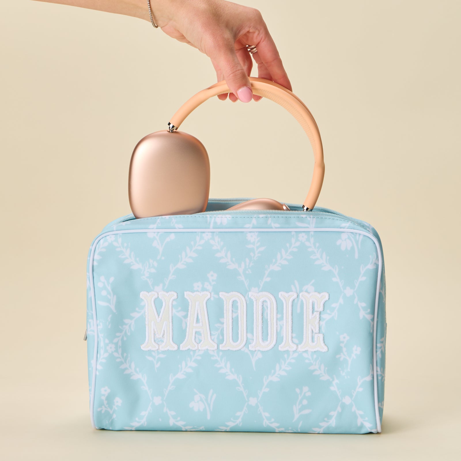 A blue floral trellis patterned toiletry bag with the name "MADDIE" in white applique, held by a hand with headphones inside.