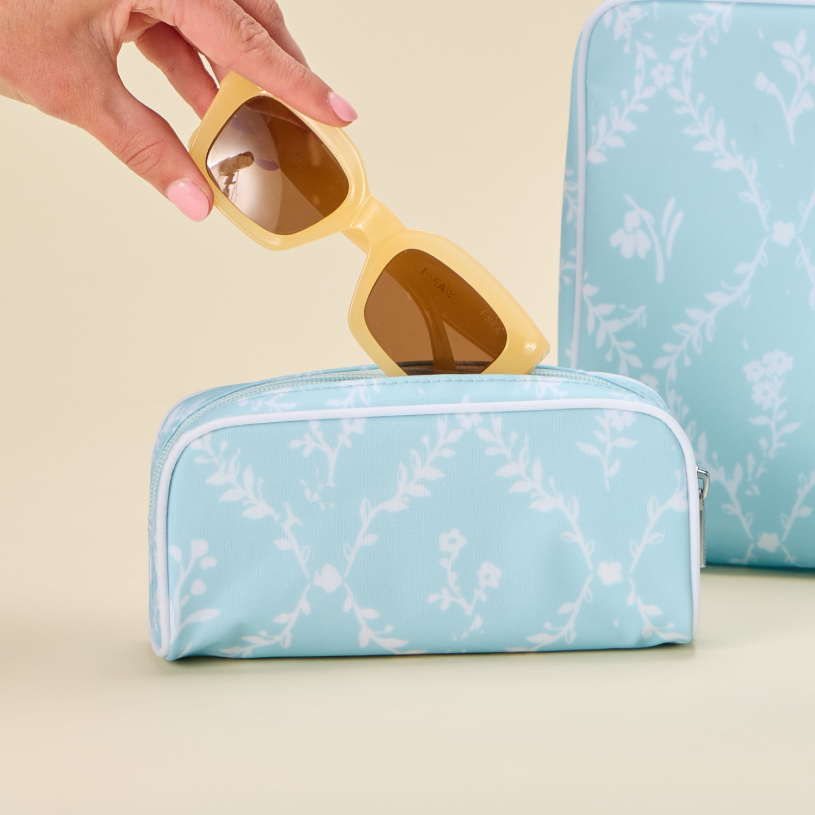 Hand placing a pair of yellow-framed sunglasses into a blue floral trellis patterned small toiletry bag.