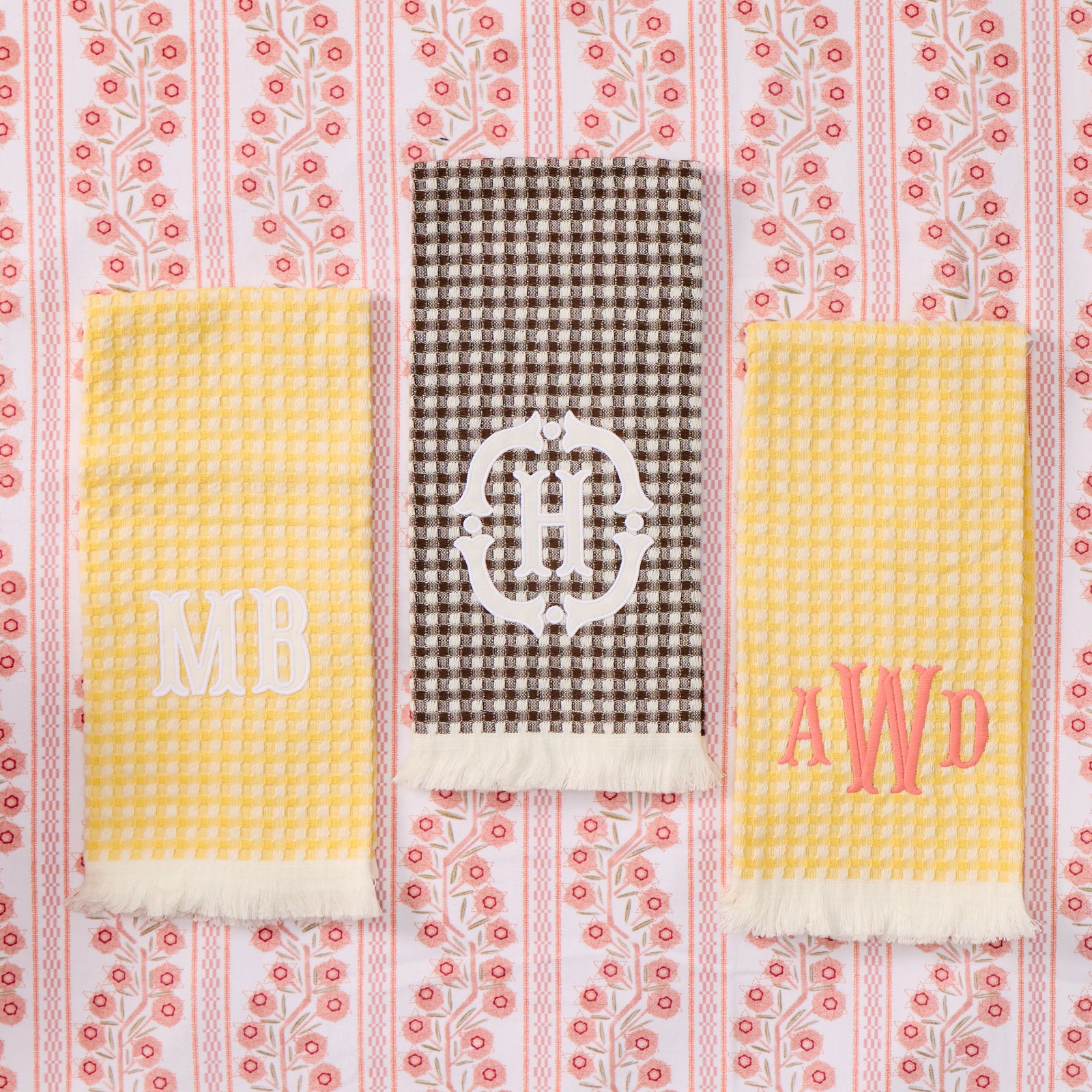Three personalized kitchen towels laid flat on a pink and white floral patterned fabric background. The left towel is pale yellow waffle-weave with white monogram letters 'MB'.