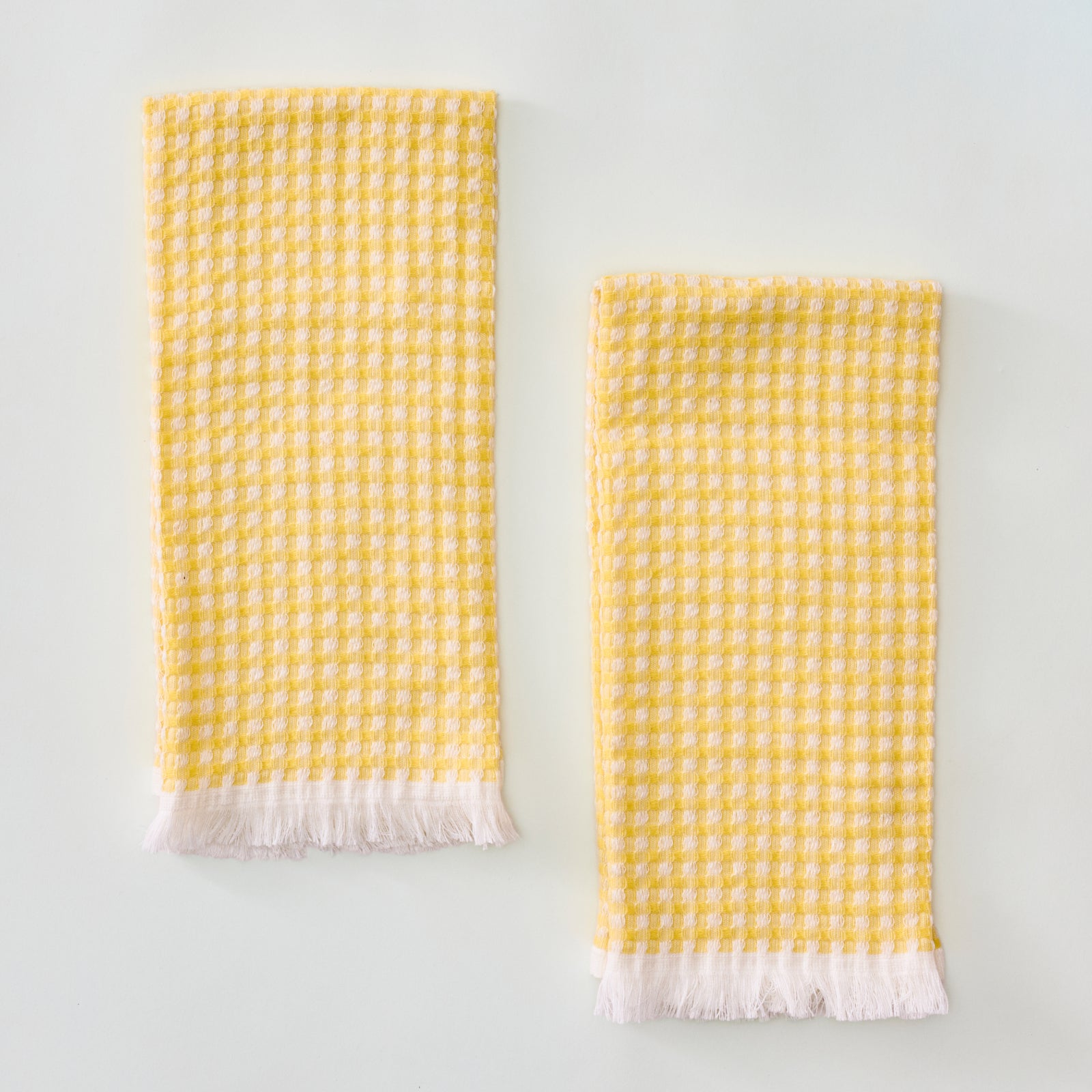 Two yellow and white checkered waffle-weave kitchen towels with white fringed hems, laid flat side-by-side against a plain light background.