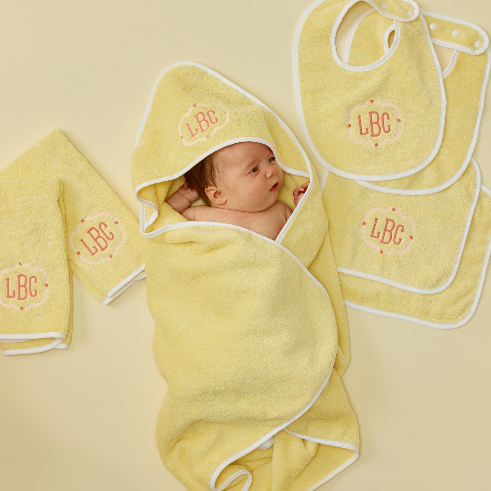 Baby wrapped in a yellow hooded towel with matching bibs, burp cloths, and washcloths featuring LBC embroidery.