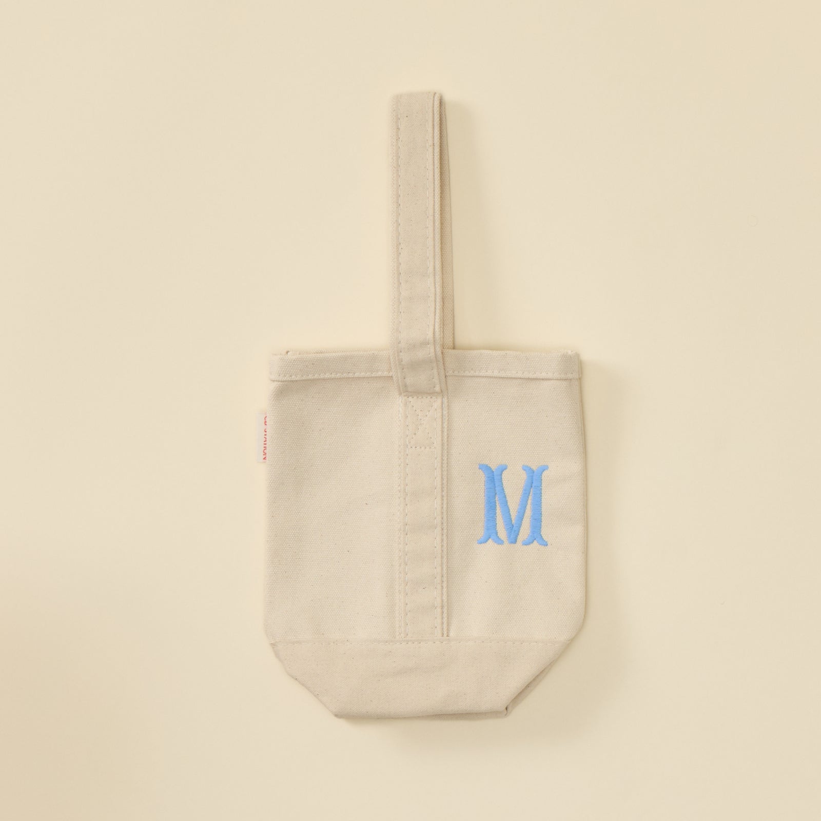 Cream canvas wine tote bag with a single strap and light blue embroidered letter M.