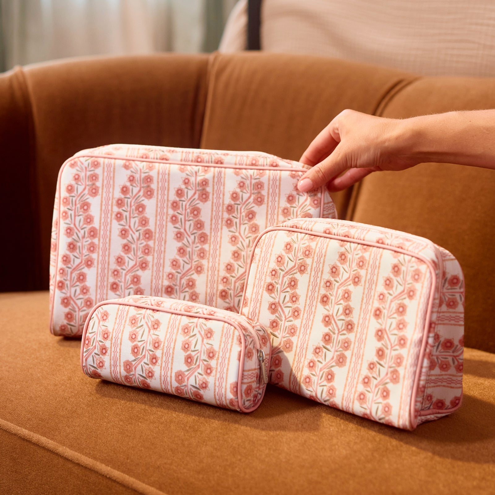 A set of three toiletry bags in small, medium, and large sizes arranged on a warm brown sofa, with a hand reaching toward the largest bag. The bags feature a pink and coral floral climbing vine stripe pattern with light pink piping.