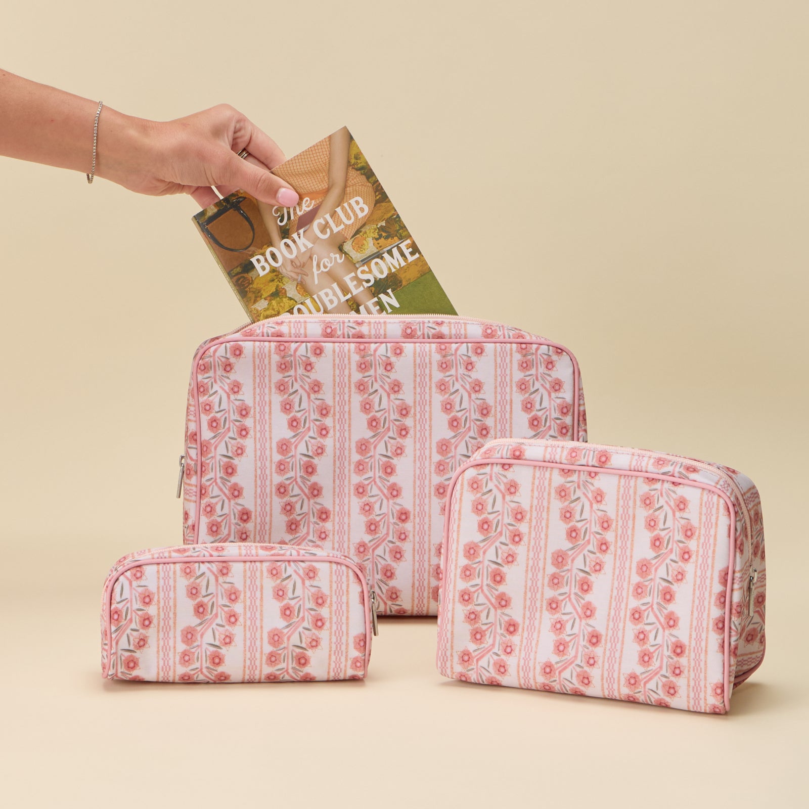 A set of three toiletry bags in small, medium, and large sizes featuring a pink and coral floral climbing vine stripe pattern, arranged on a plain light background. A hand is shown placing a book into the largest bag.