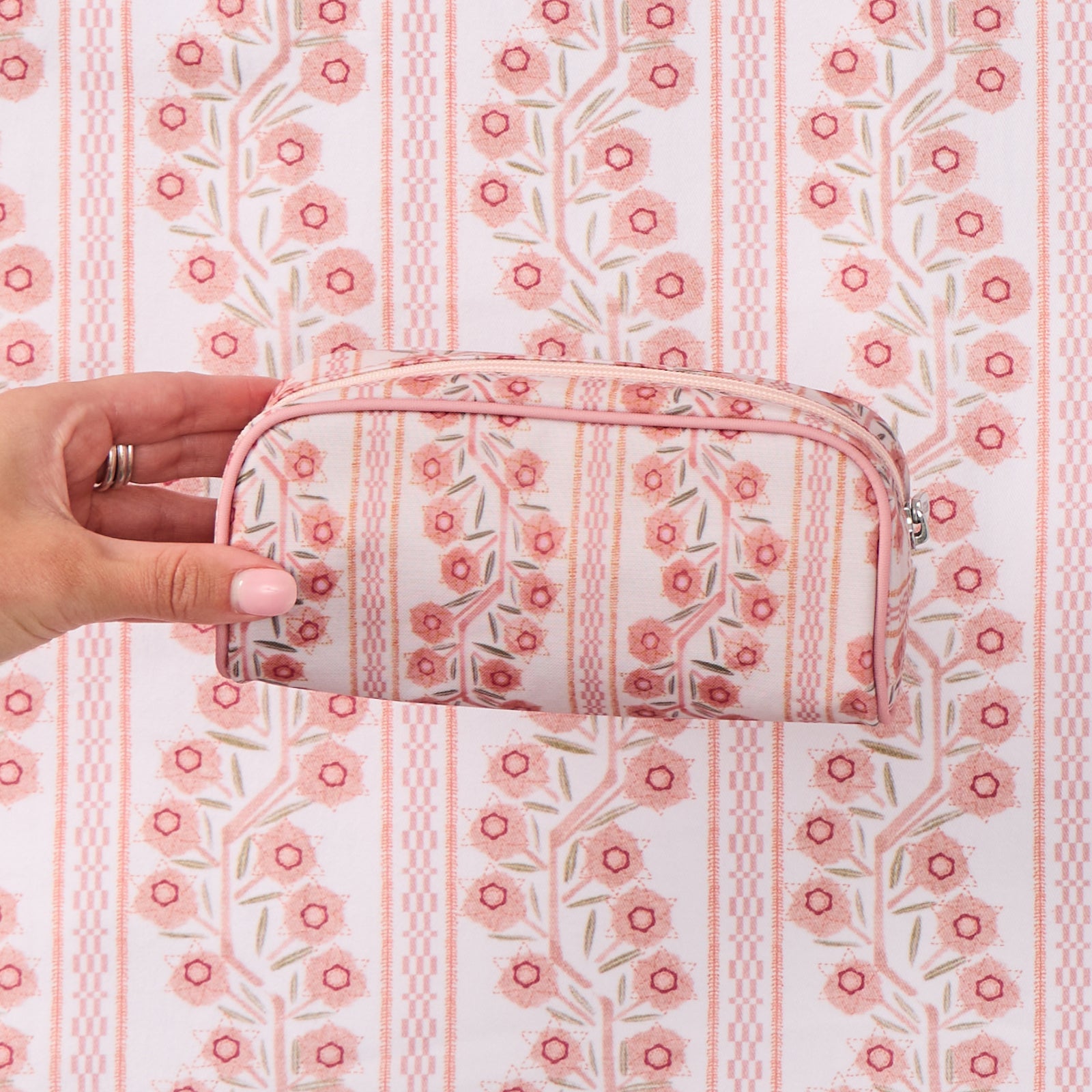 A hand holding a small toiletry bag with a pink and green floral climbing vine pattern, shown against a matching patterned background."