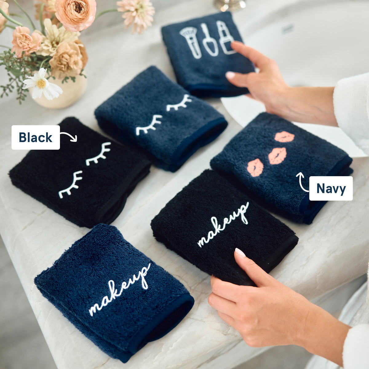 Six folded bath towels in black and navy are arranged on a light surface, each with white embroidered makeup-themed designs or words. Two hands are visible holding towels, and flowers in a vase appear in the top left.  There are "Black" and "Navy" text labels.
