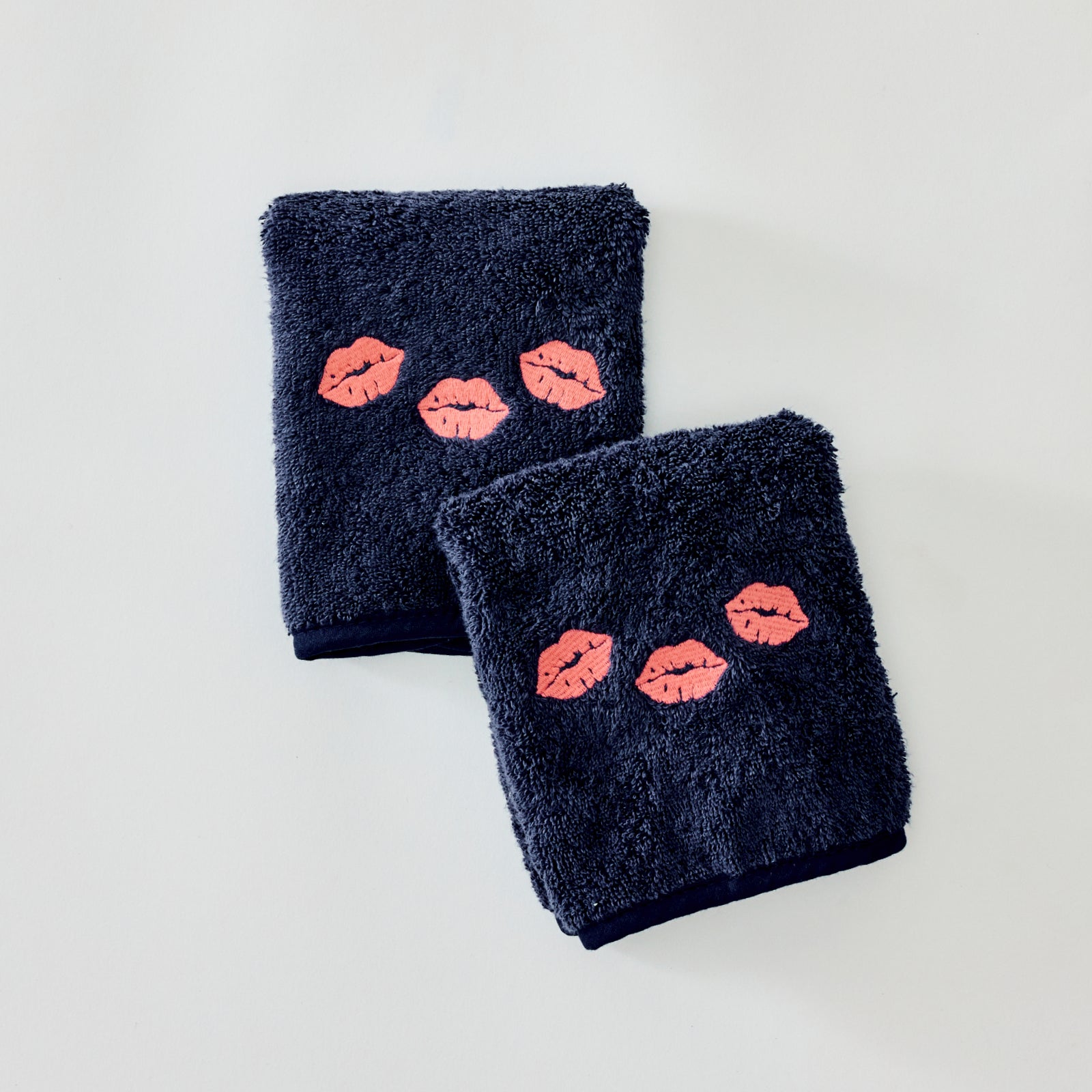 Two folded dark navy blue towels with embroidered coral pink lips, arranged on a light gray background.