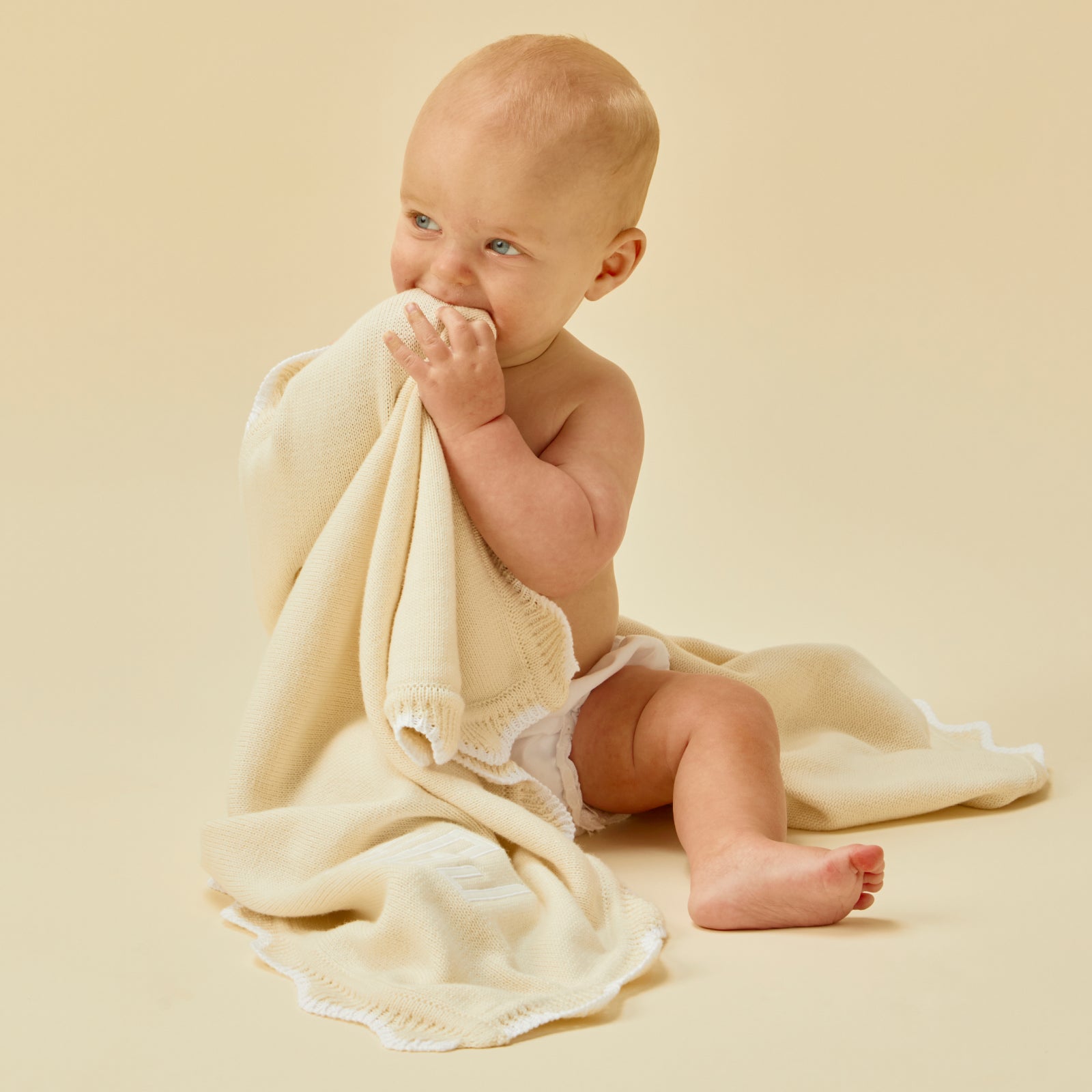 Baby holding and chewing on a soft cream baby blanket with white scalloped trim.