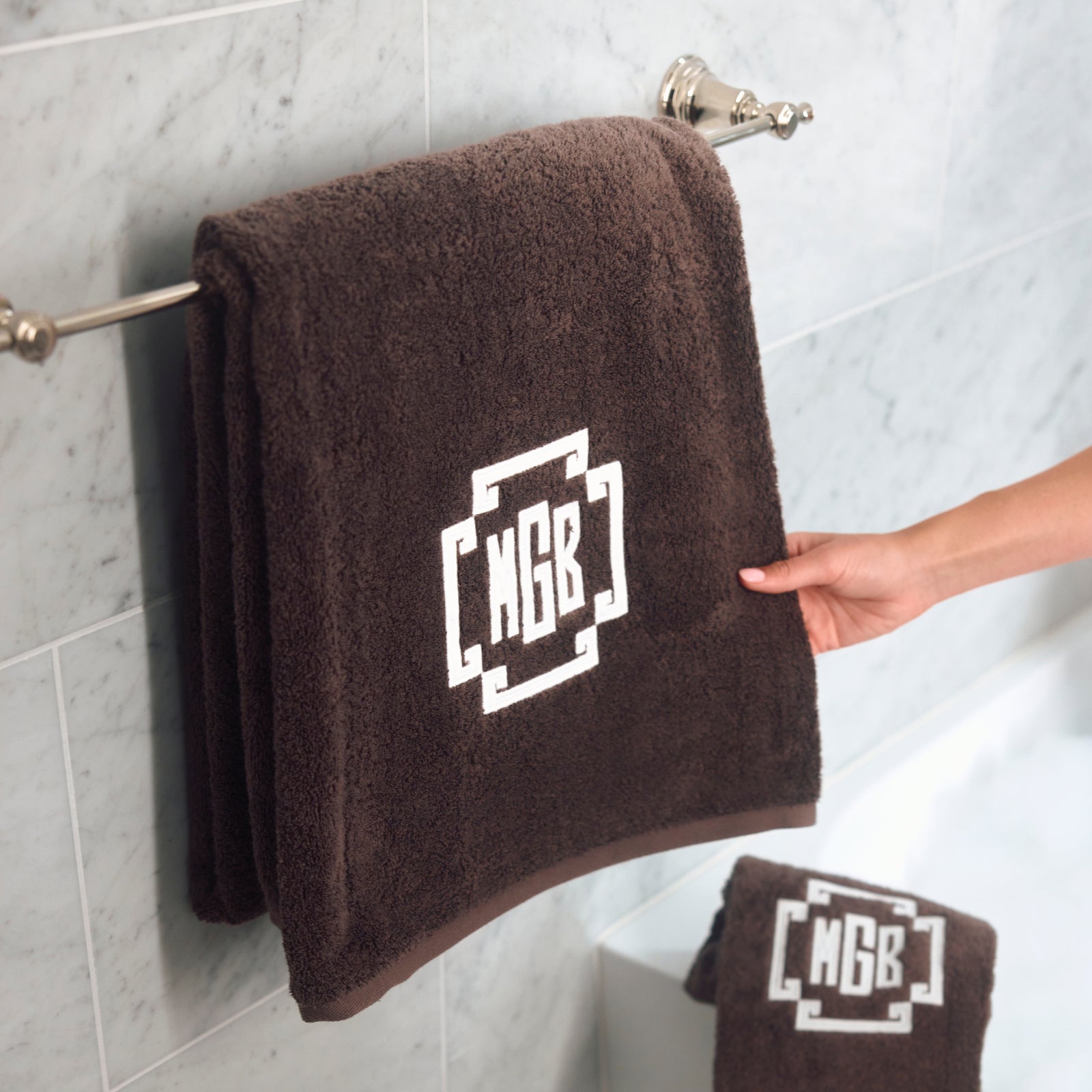 A dark brown terry cloth bath towel with a white monogram logo reading 'MGB' is hanging on a silver metal towel bar in a bathroom with light gray marble tiles. A hand is holding the edge of the towel, and a smaller matching towel is visible hanging below.