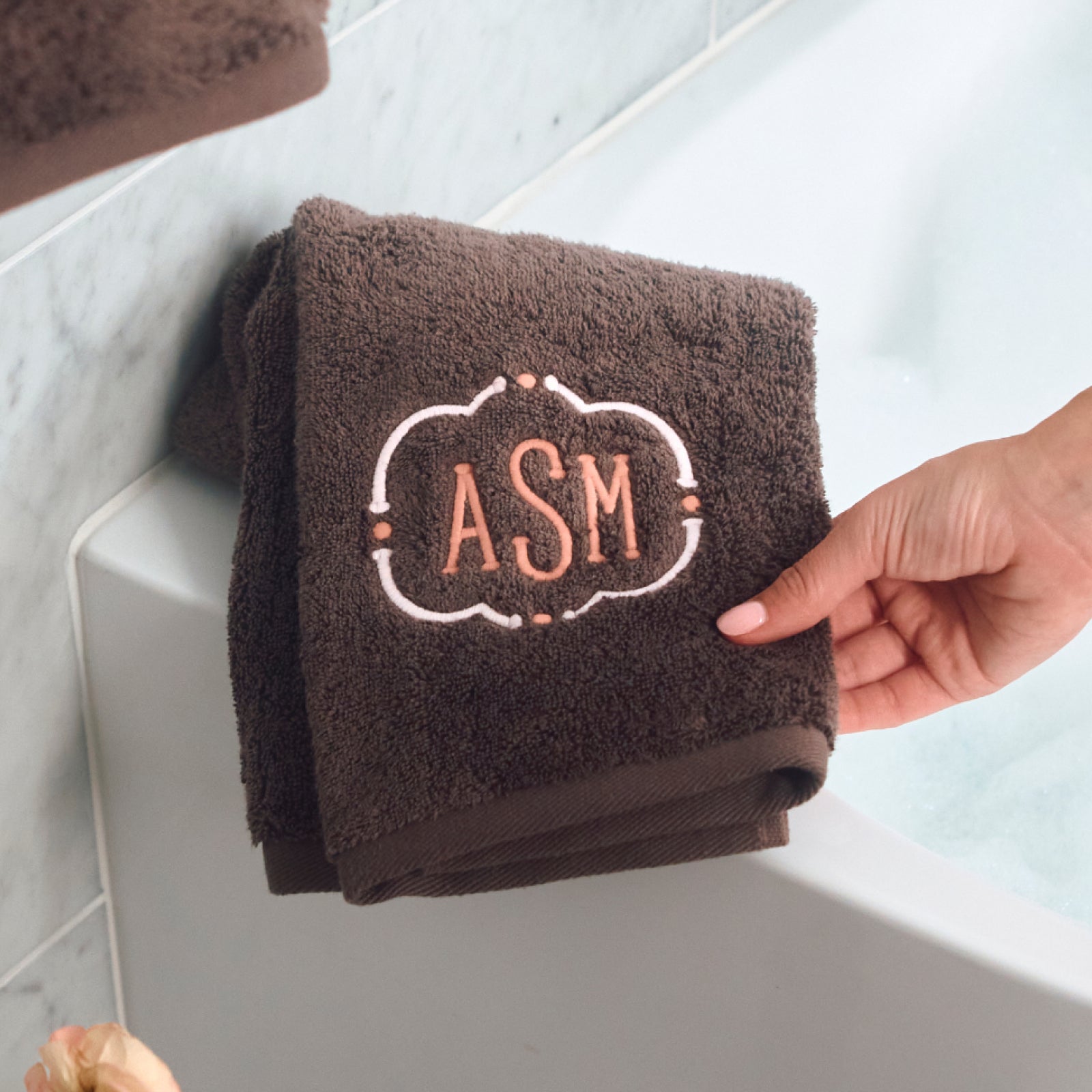 A folded dark brown bath towel with a white and rose gold monogram reading 'ASM' is held by a hand next to a white bathtub.