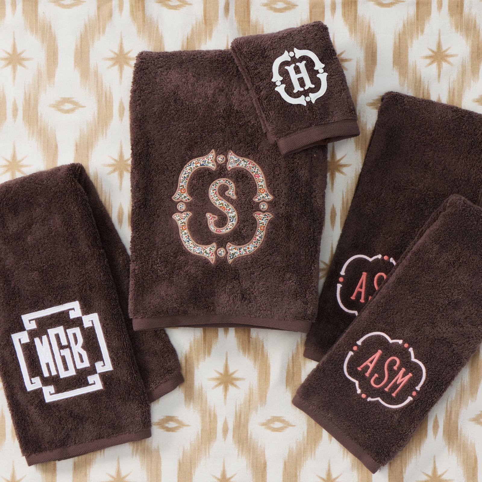 A coordinated set of dark brown plush towels and hand towels laid flat on a beige and tan patterned fabric background. Each towel has a decorative embroidered monogram: a large central ornate 'S', a small top-right 'H', a lower-left 'NGB', and a lower-right 'ASM'.