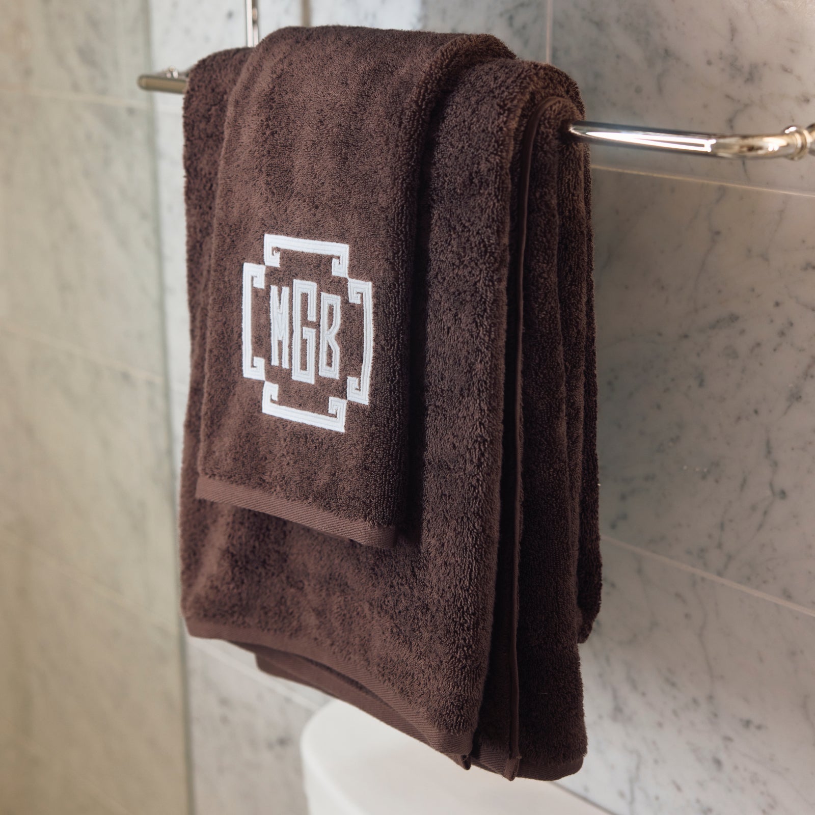 A dark brown terry cloth towel set hanging on a chrome bathroom towel bar, featuring a white embroidered geometric monogram logo that reads 'MGB'.