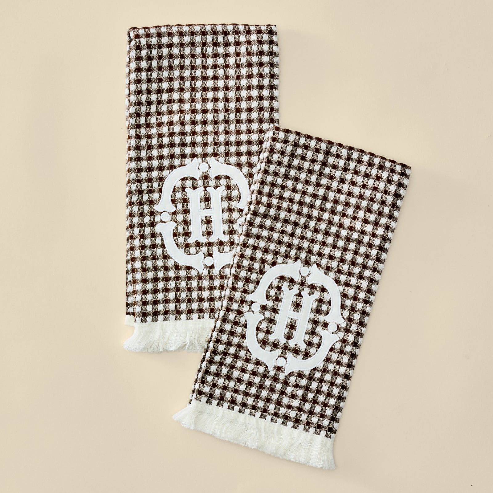 Two folded brown and white gingham check scarves with white fringe and a large white ornate monogram 'H' on each, displayed on a light beige background.