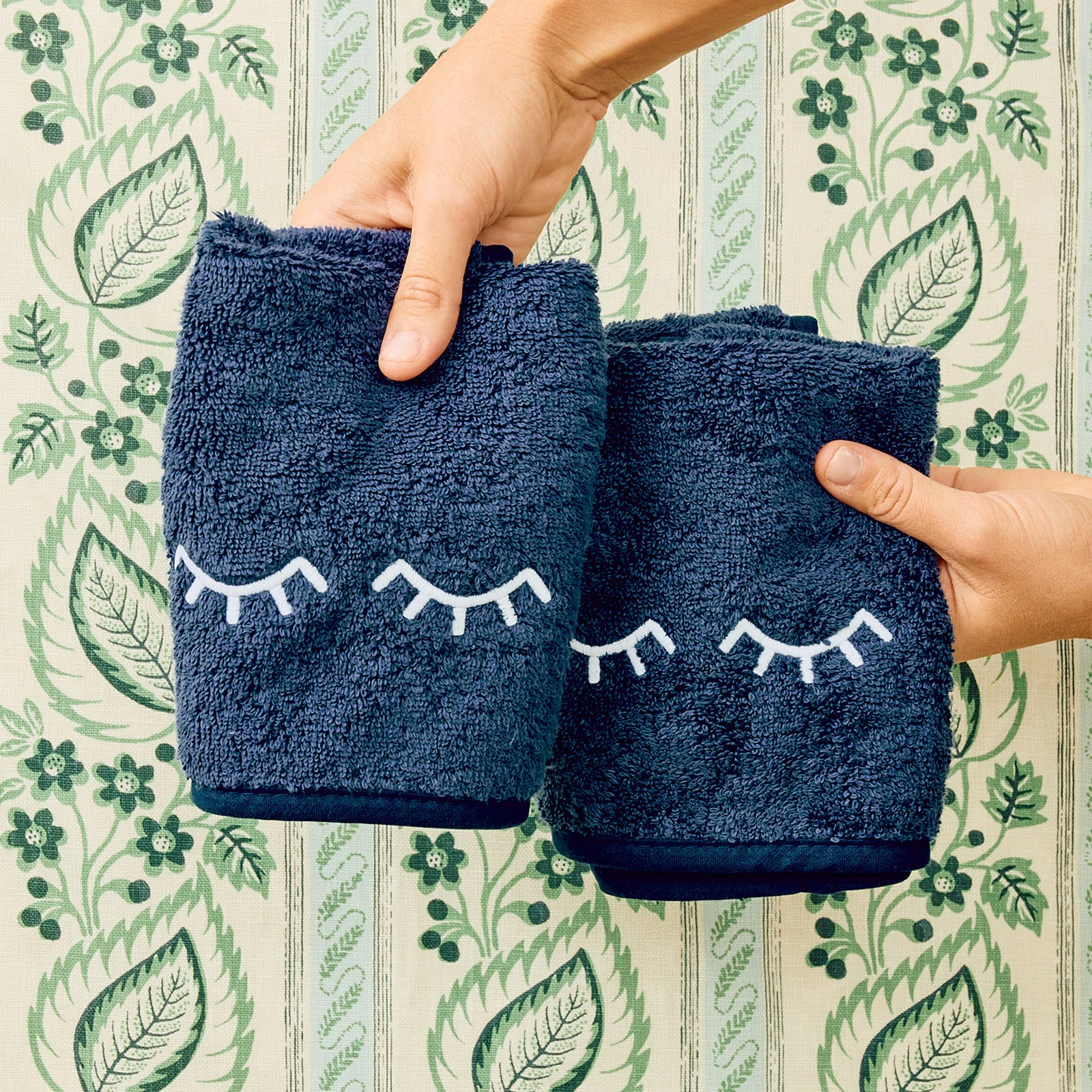 Two navy Eyelids makeup towels with white eyelash embroidery, held against a green botanical wallpaper