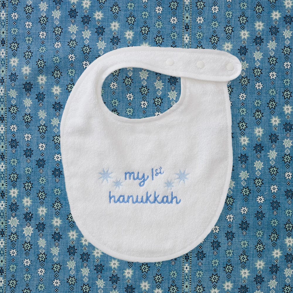 My First Hanukkah bib with blue embroidery, flat on a blue print