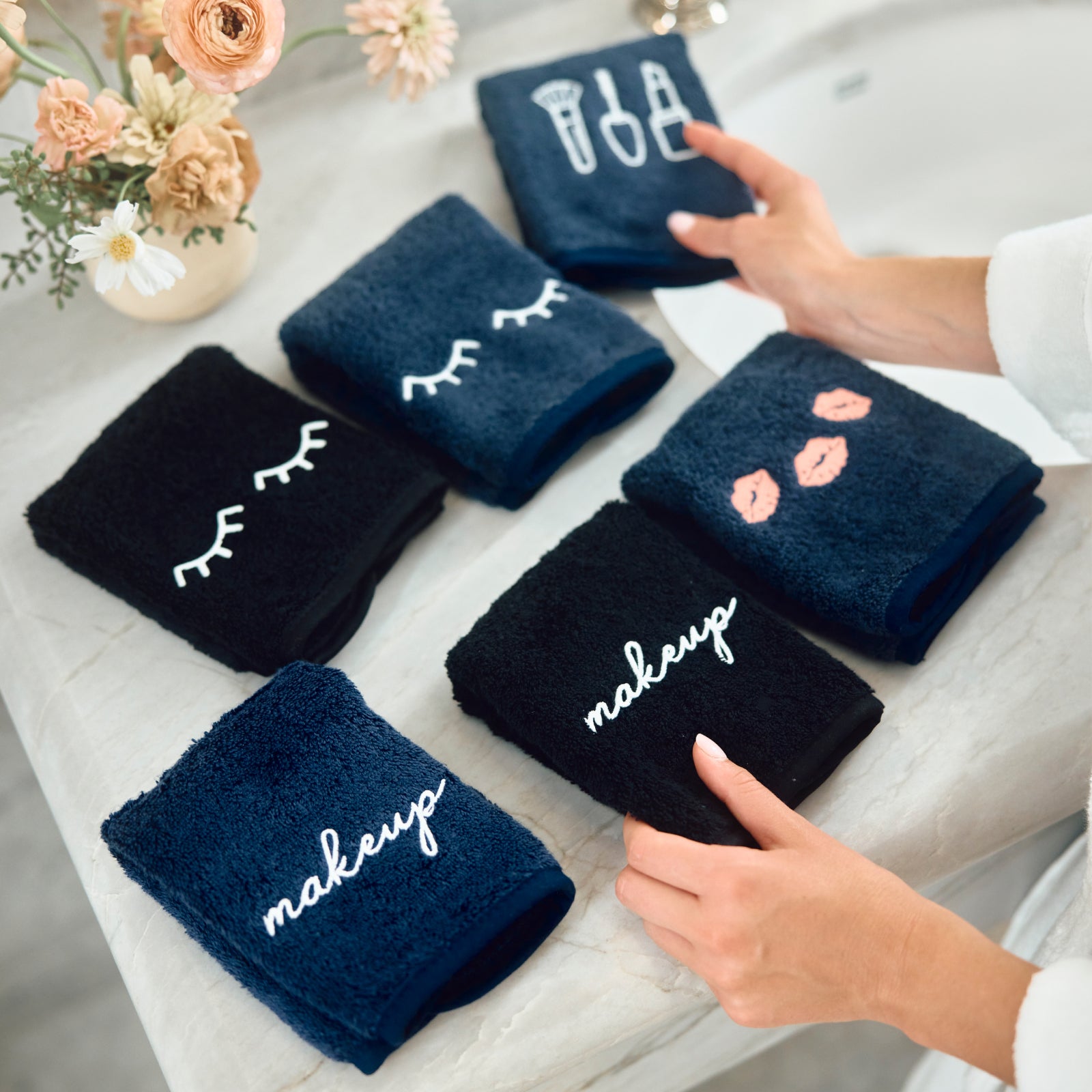 Six folded dark blue and black towels with white and pink embroidered designs are arranged on a light gray surface. Two hands are visible holding towels, with a vase of flowers in the background.