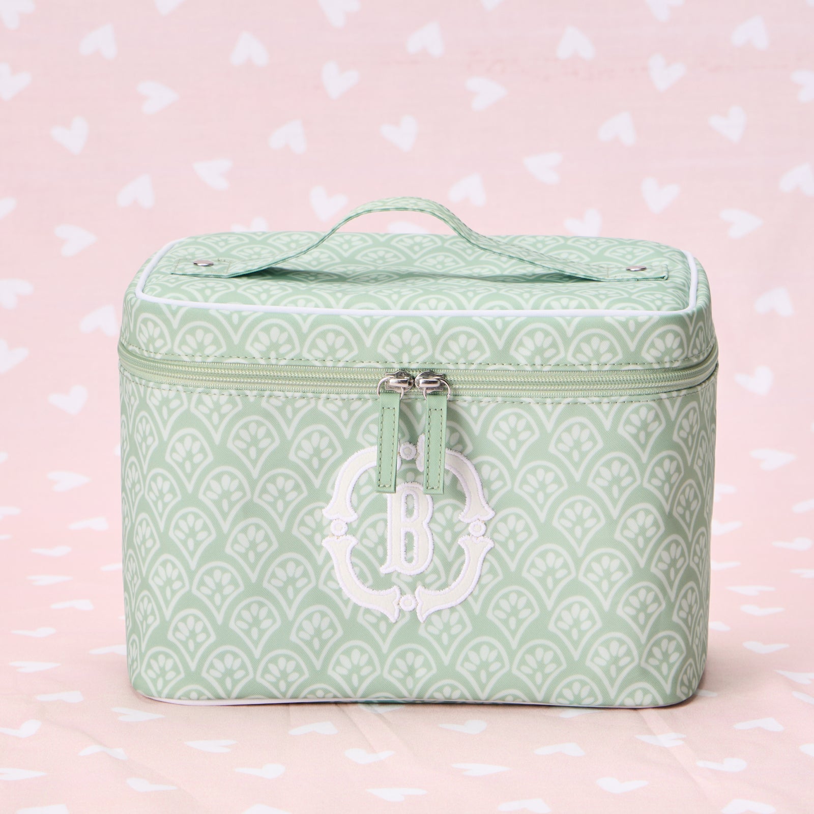A green block print vanity case featuring a white fan-patterned design and a custom white embroidered monogram 'B' on the front. The case is shown from a front-facing, straight-on view against a light pink heart-patterned background.