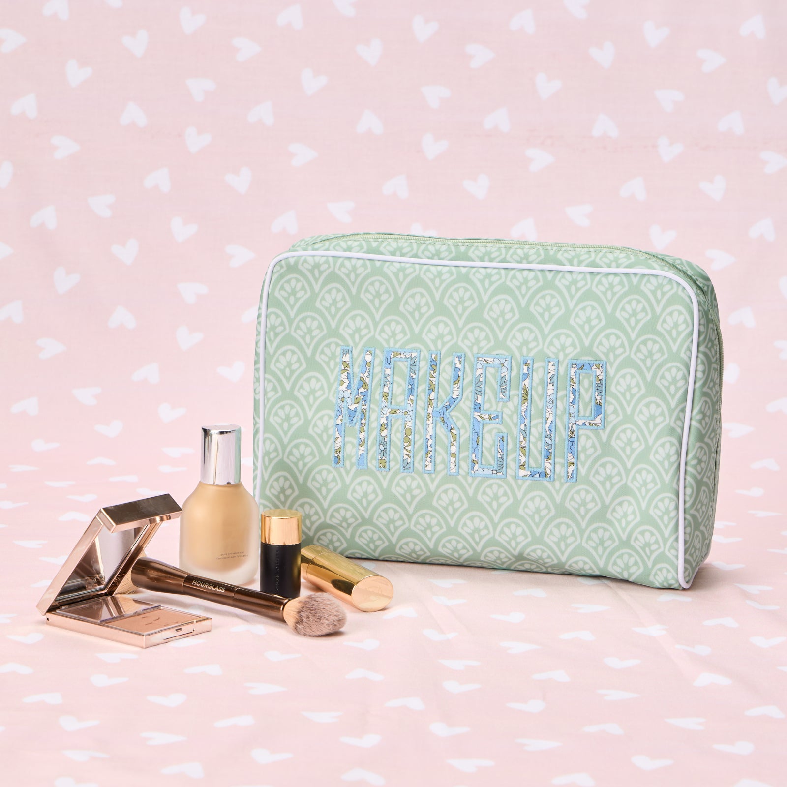 A green block-printed toiletry bag with the word "MAKEUP" in floral applique, surrounded by beauty products.