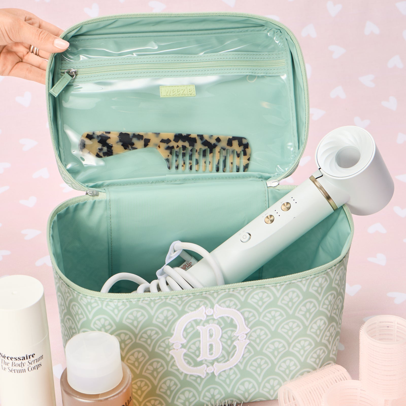 A close-up view of the interior lid of a green block print vanity case with a white fan-patterned design, featuring a transparent zippered pocket with a small 'Weezie' brand label. A tortoise-shell pattern hair comb is tucked into the pocket, and a white hair dryer is placed inside the base of the case.