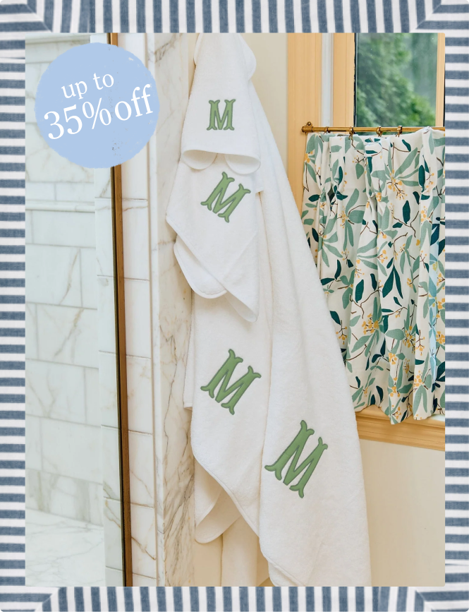 A white plush bath towel with green embroidered monogram letters hangs in a bathroom next to a floral shower curtain. A blue circular promotional sticker reads 'Up to 35% off'.
