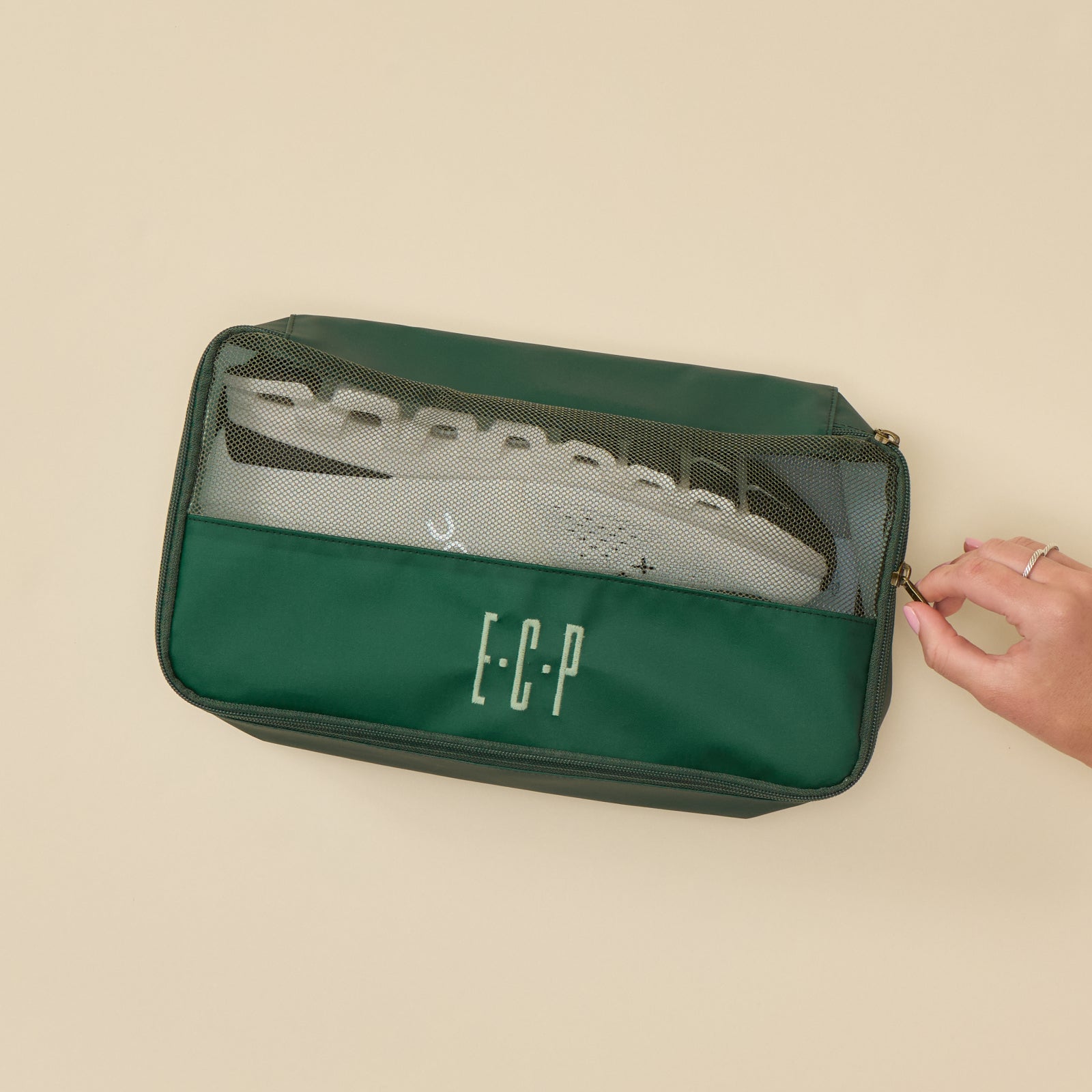 Close-up, top-down view of a rectangular, zippered Medium Packing Cube in "Hunter Green." The cube features a hunter green lower section and a matching green-trimmed clear mesh top. A pair of white sneakers is visible inside through the mesh, and a hand is shown pulling the zipper. The lower front panel is embroidered with the initials "ECP" in a light, sage-green thread.