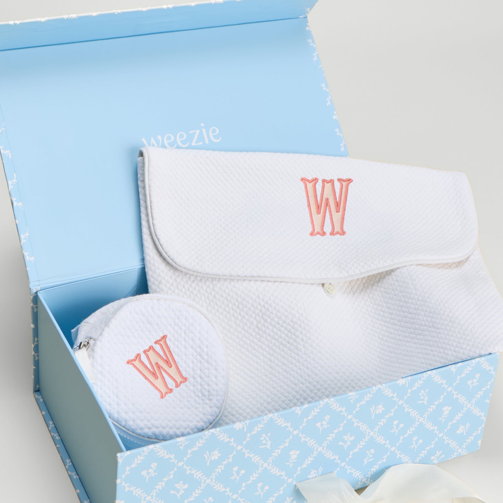 A baby gift set from Weezie in a light blue patterned box, featuring a white textured muslin pouch with a coral embroidered 'W' and a matching round zip pouch.