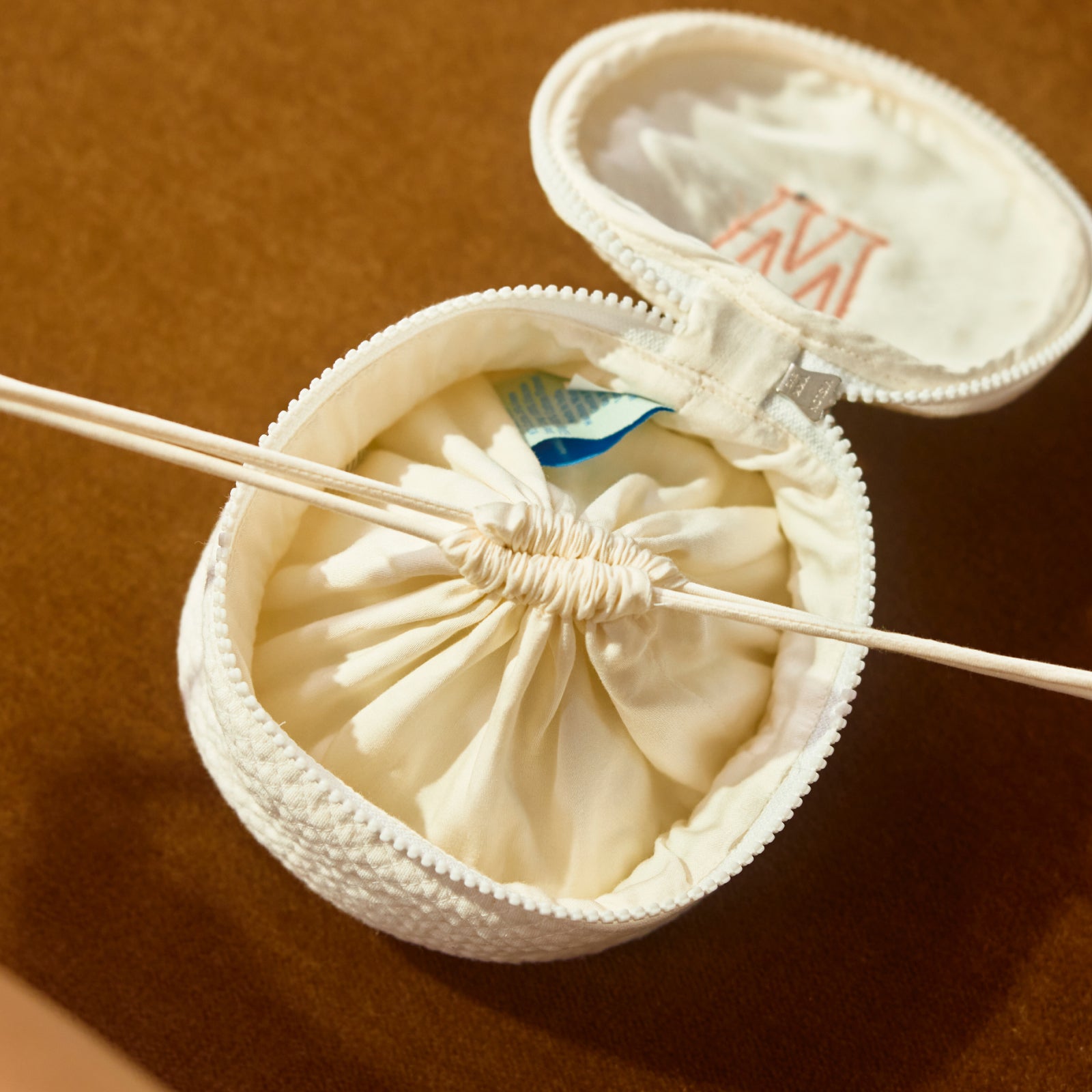 An open round white fabric pouch with a drawstring closure, shown on a warm brown background. A thin wooden stick or tool rests across the opening, and a small blue-and-white packet is visible inside.