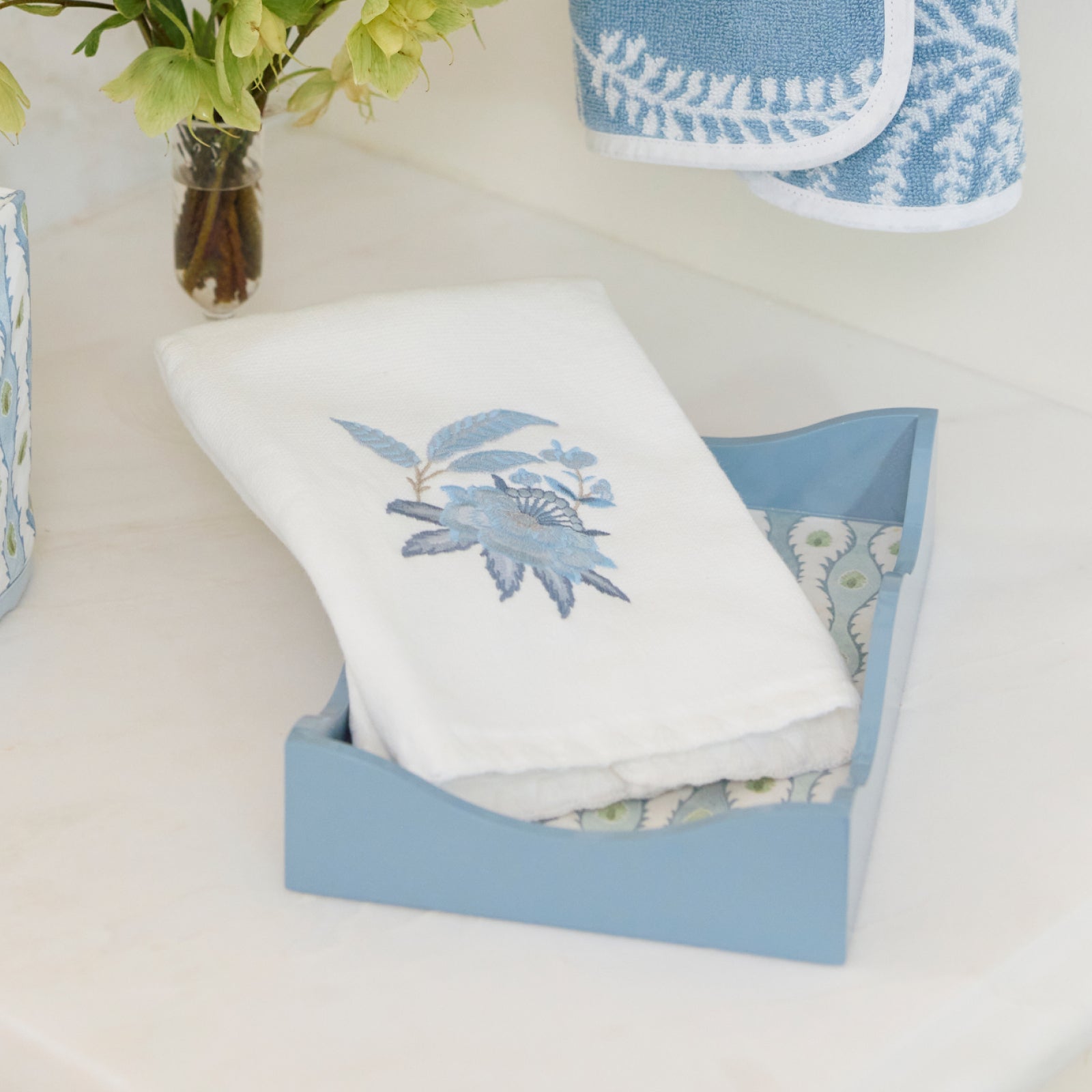 Light blue lacquer tray holding a folded white hand towel with blue floral embroidery.