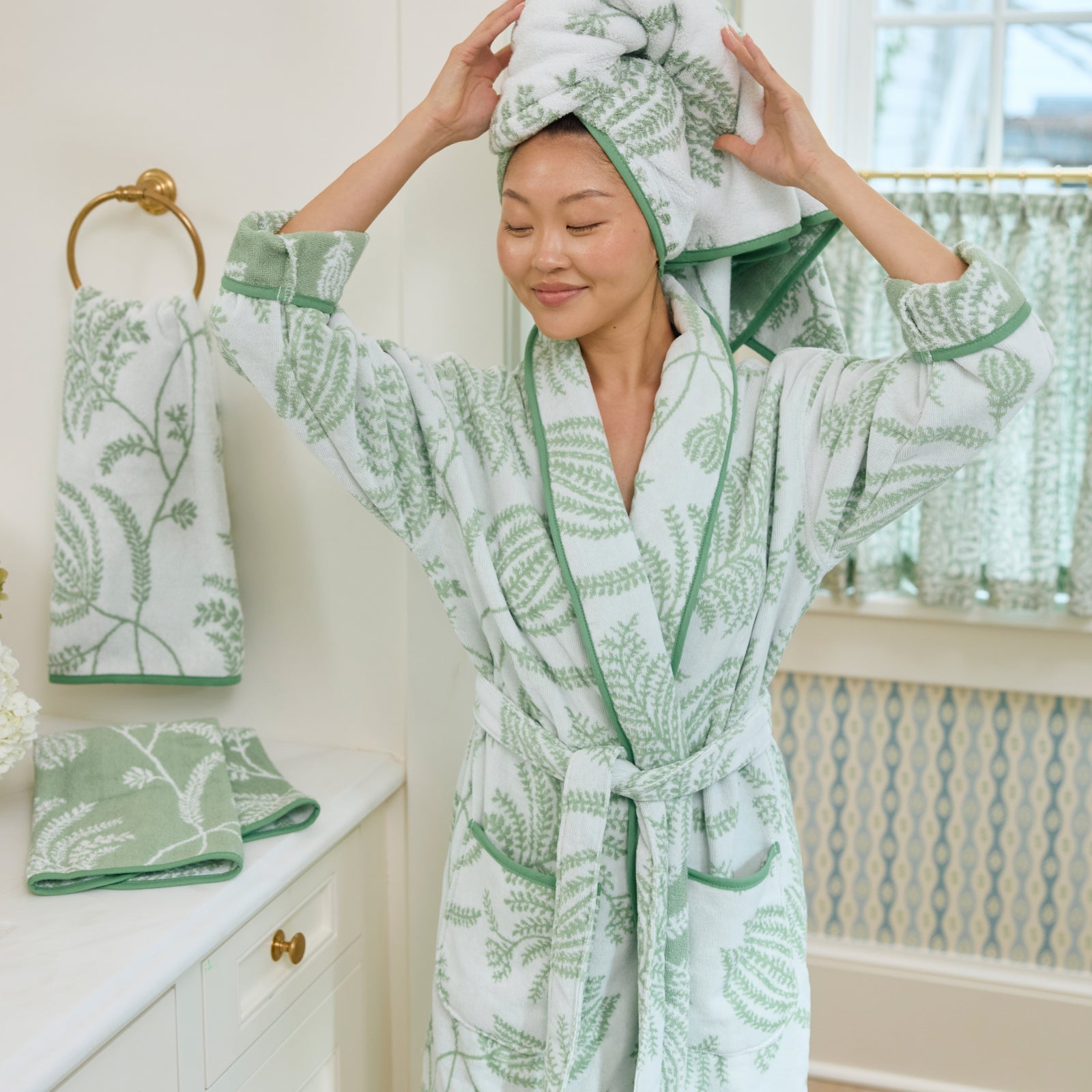 Woman wearing a long plush robe in a sage green and white floral print with green piped edges.