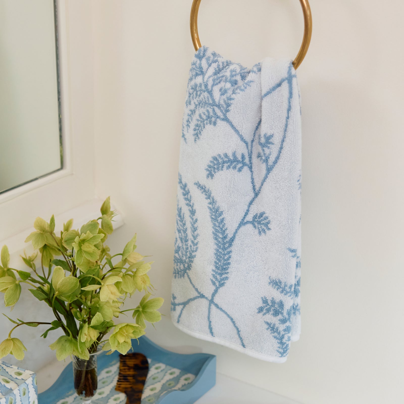 White towel with sky blue Palmero floral pattern hanging on a brass ring next to flowers.