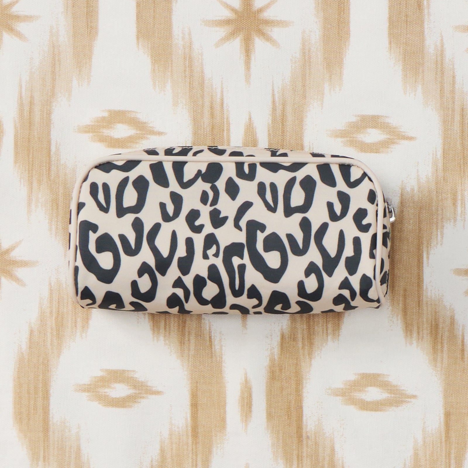 A small zippered pouch with a black leopard print pattern on a cream background, placed on a cream and tan ikat patterned fabric surface.