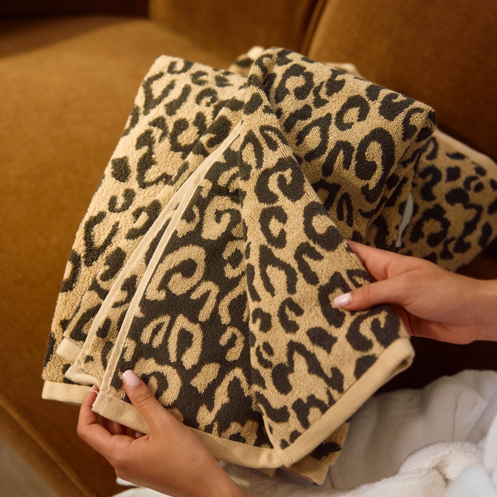 A close-up of a leopard-print towel being held by two hands, with a matching brown sofa in the background.