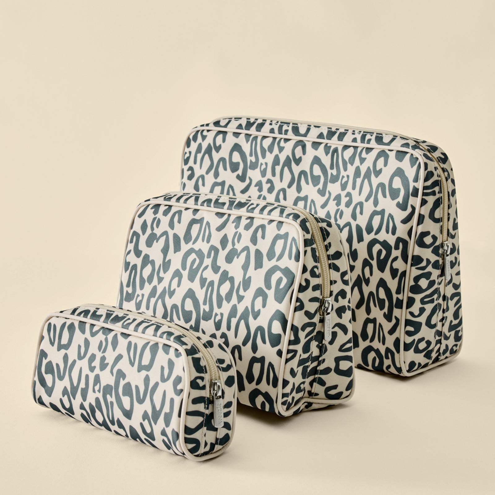 Three leopard-print cosmetic bags in different sizes arranged on a warm beige background.